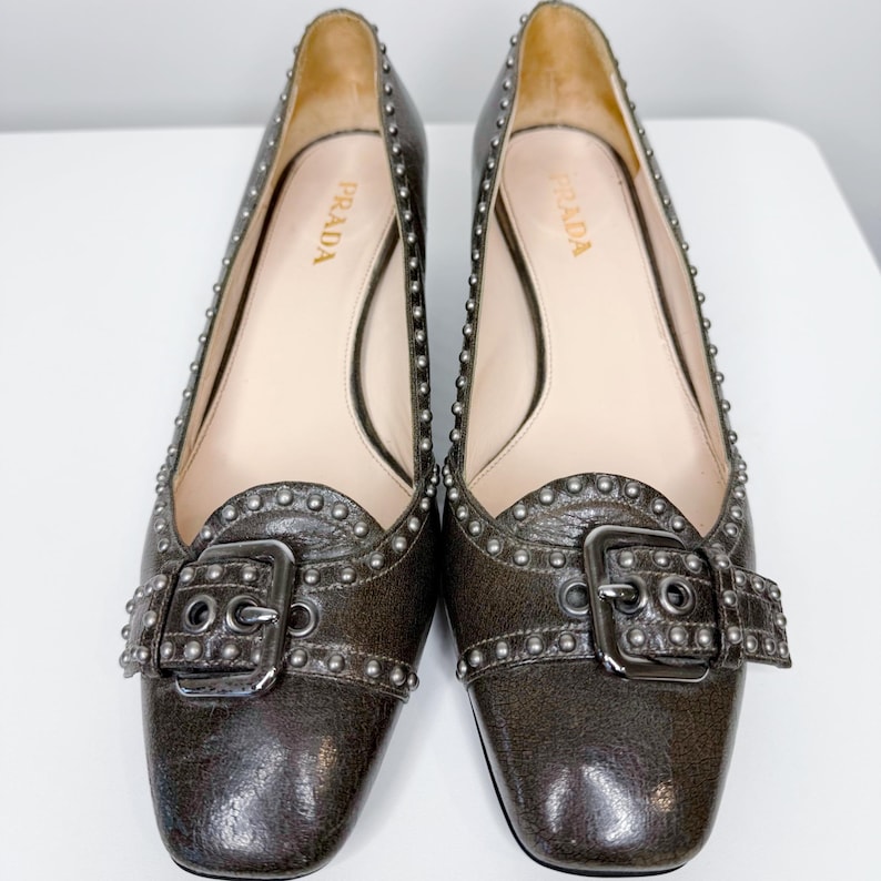 May include: A pair of dark brown leather flats with silver-tone stud accents and buckle closures. The shoes have a square toe and the brand name "PRADA" is visible on the insole. The flats are displayed on a white surface.
