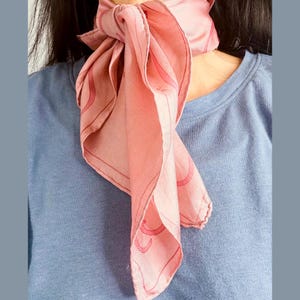 Puede incluir: Un pañuelo de seda rosa claro atado al cuello. El pañuelo presenta un patrón repetitivo de líneas curvas en un tono más oscuro de rosa. El fondo es una camisa azul claro.