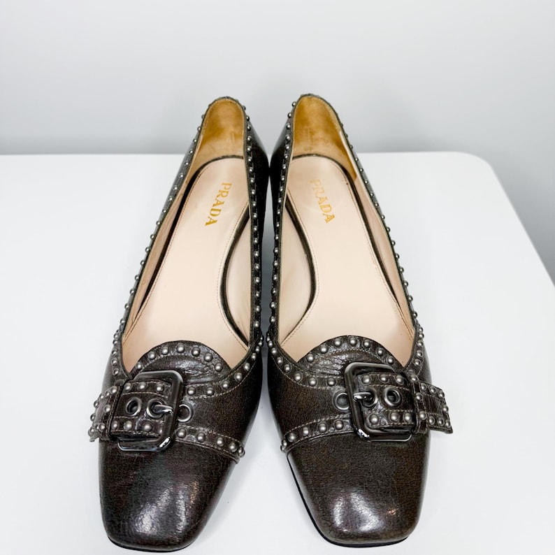 May include: A pair of brown leather Prada flats with silver-tone studding and buckle details. The shoes have a square toe and a low heel. The Prada logo is visible on the insole.