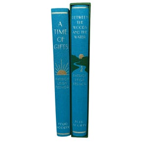 Può includere: Due libri con copertina rigida con copertine color teal. Il libro a sinistra è intitolato "A Time of Gifts" con un disegno a forma di raggio di sole dorato. Il libro a destra è intitolato "Between the Woods and the Water" con un'illustrazione di fiume.