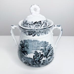 May include: White ceramic sugar bowl with a lid, decorated with a black and white rural landscape and floral patterns. The bowl has two handles and a decorative knob. A classic piece for serving sugar or other condiments.