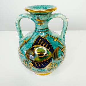May include: A turquoise ceramic vase with two handles and a gold rim. The vase is decorated with a fish design in navy blue, green, and gold accents. The vase has a round shape and a textured surface.