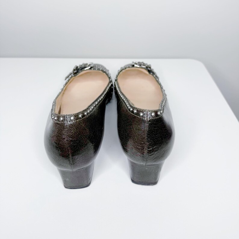May include: A pair of brown leather shoes with a low heel. The shoes have a decorative buckle and are studded with silver-colored metal studs around the top edge. The shoes are displayed on a white surface.