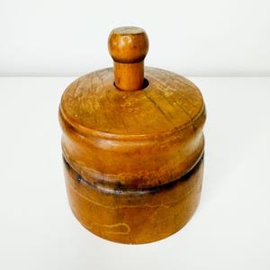 Peut inclure: Un presse-beurre cylindrique en bois avec une finition brun clair naturelle et des motifs de grain de bois visibles. L'objet se compose d'une base circulaire et d'un couvercle assorti avec une poignée centrale verticale.