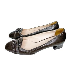 May include: A pair of brown leather flats with a small heel. The shoes feature a buckle detail on the vamp and are adorned with silver-colored studs along the edges. The interior is a light beige color.