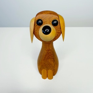 May include: A wooden dog figurine with a light brown body, head, and ears. The dog has large black eyes and a black nose. The figurine is sitting upright with its small legs visible. The wood grain is visible.