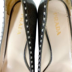 May include: Close-up of a pair of Prada high heels. The shoes are dark gray with silver studs along the edges. The insole is cream-colored with the Prada logo in gold lettering.