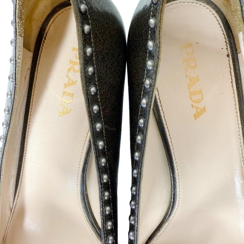 May include: Close-up of a pair of Prada high heels. The shoes are dark gray with silver studs along the edges. The insole is cream-colored with the Prada logo in gold lettering.