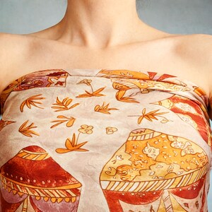 May include: A strapless scarf or fabric wrap featuring a light beige background with a repeating pattern of orange and red stylized mushroom shapes, interspersed with small orange floral and leaf motifs across the textured material surface.