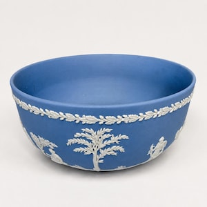 May include: A matte blue ceramic bowl featuring white relief decorations of neoclassical figures and trees around the exterior. A continuous white laurel leaf garland border encircles the upper rim of the bowl, providing a detailed contrast.
