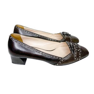 May include: A pair of dark brown leather flats with silver-tone stud accents and buckle details. The shoes have a low heel and a pointed toe. The interior is a light beige color.