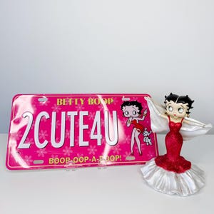 May include: A pink license plate with the text "2 CUTE 4U" and a Betty Boop graphic. A Betty Boop figurine in a red dress and white cape stands to the right. The license plate has the text "BOOP-OOP-A-DOOP!"
