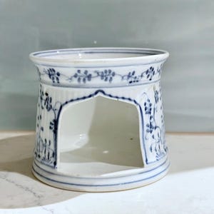 Puede incluir: Un portavelas de cerámica blanca con diseños florales azules. El soporte tiene forma cilíndrica con una abertura arqueada y un borde decorativo. El diseño azul incluye hojas y vides.