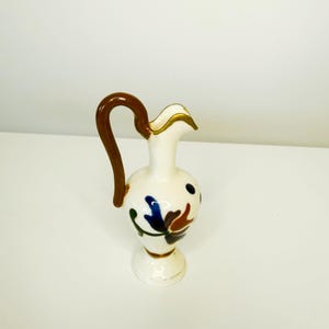 May include: A white ceramic jug with a brown handle and gold trim. The jug is decorated with blue, green, and brown floral designs. The jug stands on a small, round base.
