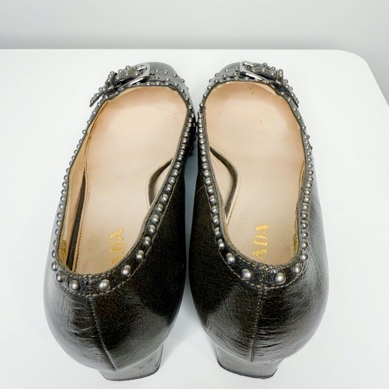 May include: Pair of dark gray leather flats with silver-tone studding around the edges and a buckle detail. The interior is a light beige color with the word "PRADA" printed in gold.