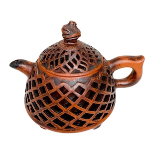 Op de afbeelding: Een terracotta theepot met een traliewerkontwerp. De theepot heeft een afgeronde body, een gebogen handvat en een tuit. Het deksel is bekroond met een klein, gebeeldhouwd figuur. De theepot is waarschijnlijk handgemaakt en heeft een rustieke uitstraling.