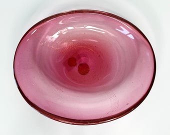 May include: A round, pink glass bowl with a deep center. The bowl has a gradient of color, transitioning from a lighter pink at the edges to a darker, reddish-pink in the center. The rim has a darker, textured finish.