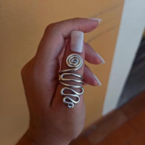 May include: A silver-colored wire ring with a spiral design on the top and a wavy pattern down the finger. The ring is worn on a finger, showcasing its unique, artistic shape. The ring's design is eye-catching and modern.