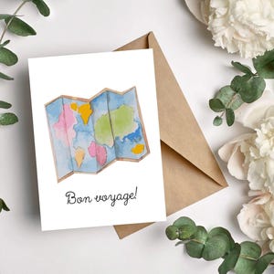 May include: A white greeting card with a watercolor map design in blue, pink, yellow, and green, with the phrase "Bon voyage!" in black script. A brown envelope is behind the card, with white flowers and green leaves in the background.