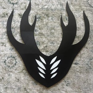 May include: A black, shield-shaped wall decoration featuring a symmetrical, flame-like silhouette with four upward-curving points on each side. The center of the piece contains a vertical column of eight white, leaf-shaped cutouts arranged in pairs.