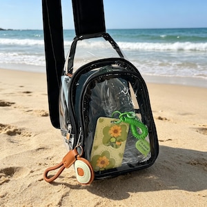 Op de afbeelding: Transparante crossbodytas met zwarte rand, een zwarte band en een bruine leren sleutelhanger. In de tas zit een telefoonhoesje met een zonnebloemontwerp en een groene sleutelhanger met de tekst "I'm stuck on you!"