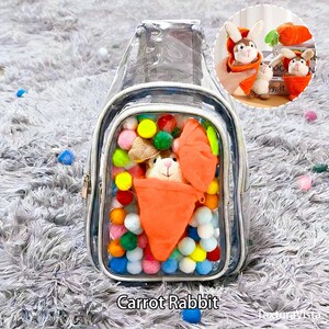 Puede incluir: Mochila transparente con ribete gris, llena de pompones coloridos y un conejo de peluche con un disfraz de zanahoria naranja. El texto "Carrot Rabbit" est&aacute; en la parte inferior de la imagen. Se muestran conejos de peluche adicionales con disfraces de zanahoria.