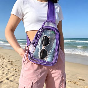 May include: A clear, purple sling bag with a water bottle and sunglasses inside. The bag has a purple strap and a small charm. The bag is worn over the shoulder. The background is a beach with sand and ocean.
