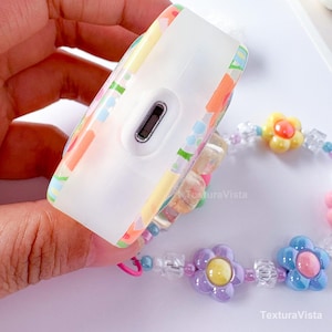 K&ouml;nnte beinhalten: Eine wei&szlig;e, rechteckige H&uuml;lle mit einem farbenfrohen Blumenmuster. Die H&uuml;lle hat einen Ladeanschluss und ist mit einem Perlenarmband mit Blumen- und Kristallakzenten verbunden. Das Armband zeigt gelbe, lila und blaue Blumen.