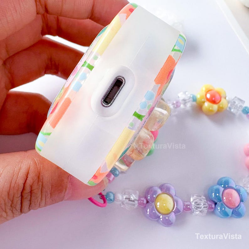 K&ouml;nnte beinhalten: Eine wei&szlig;e, rechteckige H&uuml;lle mit einem farbenfrohen Blumenmuster. Die H&uuml;lle hat einen Ladeanschluss und ist mit einem Perlenarmband mit Blumen- und Kristallakzenten verbunden. Das Armband zeigt gelbe, lila und blaue Blumen.