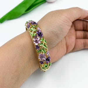 Pode incluir: Pulseira dourada articulada adornada com flores roxas e lavanda e folhas verdes. O design floral é colocado sobre um fundo de filigrana dourada. A pulseira é uma joia.