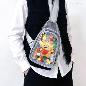 Puede incluir: Bolso bandolera transparente con una correa reflectante. La bolsa contiene un juguete de peluche amarillo con forma de perro que lleva un coraz&oacute;n rojo, rodeado de pompones de colores. El bolso se lleva al hombro.