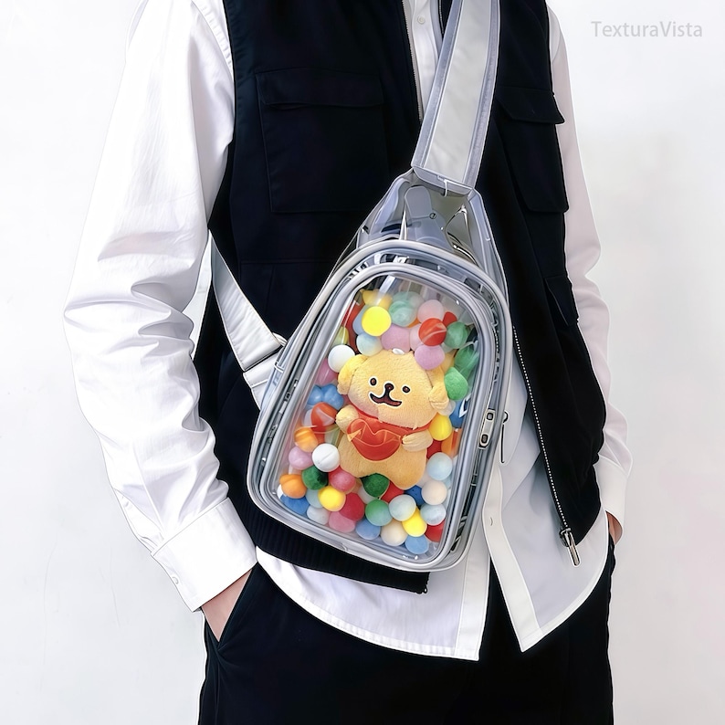 Puede incluir: Bolso bandolera transparente con una correa reflectante. La bolsa contiene un juguete de peluche amarillo con forma de perro que lleva un coraz&oacute;n rojo, rodeado de pompones de colores. El bolso se lleva al hombro.