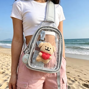 May include: Clear crossbody bag with a gray strap and trim. The bag contains a small plush dog with a red heart and a light blue umbrella. The bag is worn over the shoulder.
