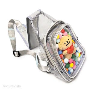 Puede incluir: Bolso bandolera transparente con correa y ribetes plateados. El bolso tiene un compartimento con cremallera lleno de pompones de colores y un peque&ntilde;o oso de peluche con una bufanda roja. El bolso est&aacute; dise&ntilde;ado para llevarse al hombro.