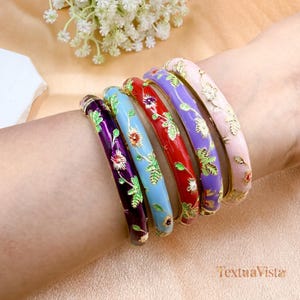 Puede incluir: Seis brazaletes de esmalte coloridos con diseños florales. Los brazaletes son en tonos morado, azul, rojo y rosa, con detalles dorados. Cada brazalete presenta un patrón floral diferente con hojas verdes.
