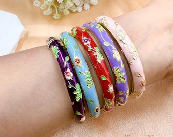 Könnte beinhalten: Sechs bunte Emaille-Armreifen mit floralen Designs. Die Armreifen sind in den Farbtönen Lila, Blau, Rot und Rosa gehalten und mit goldenen Akzenten versehen. Jeder Armreif hat ein anderes Blumenmuster mit grünen Blättern.