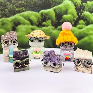May include: A collection of decorative crystal figurines, each adorned with small, round eyeglasses. The figurines are various colors and shapes, some with hats or accessories. The background is a blurred green.