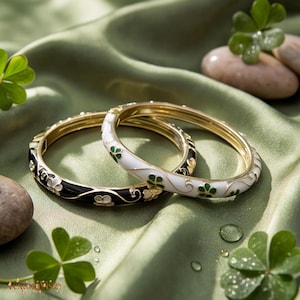 May include: Two hinged bangle bracelets, one in black and one in white, both featuring gold-tone metal edges and delicate enamel floral patterns with green and white accents. The bracelets have a smooth, polished finish and a secure clasp mechanism.