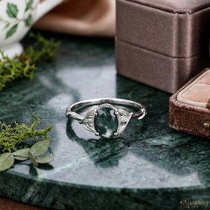 May include: A silver ring featuring an oval-shaped moss agate gemstone set in a prong setting. The band is designed with intricate leaf-like metal engravings on both sides of the central stone, creating a detailed botanical appearance.