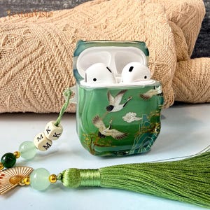 May include: Green AirPod case with a crane design. The case is open, revealing white AirPods. A green tassel and beaded charm with the letters "M K M" are attached. The case has a glossy finish.