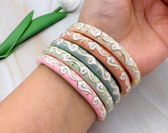 May include: A collection of colorful enamel bangles in shades of pink, peach, green, and blue, adorned with a white cloud design and gold trim. The bangles are stacked on a wrist, showcasing their decorative pattern and vibrant colors.