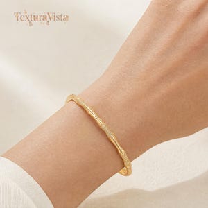 May include: A gold-toned bangle bracelet featuring a textured bamboo-inspired design with alternating smooth and granular segments. The jewelry piece encircles the wrist, showcasing a metallic finish and a repeating organic stalk pattern throughout.