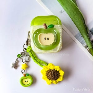K&ouml;nnte beinhalten: Gr&uuml;nes AirPod-Case im Apfel-Design mit passendem Charm. Das Case hat ein klares, strukturiertes Design mit einer gr&uuml;nen Apfelgrafik. Eine gelbe H&auml;kelsonnenblume und andere Charms h&auml;ngen an dem Case. Das Case dient zum Schutz und zur Aufwertung Ihrer AirPods.