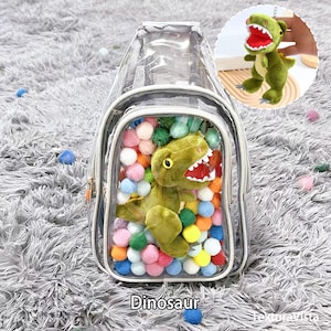 Puede incluir: Una mochila transparente con un peluche de dinosaurio y pompones de colores en su interior. La mochila tiene un ribete plateado y cierre de cremallera. Tambi&eacute;n se ve un peque&ntilde;o llavero de dinosaurio. La palabra "Dinosaurio" est&aacute; impresa en la parte inferior.