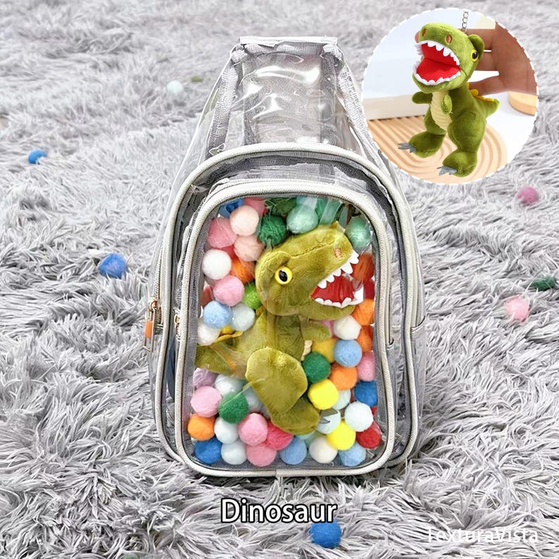 Puede incluir: Una mochila transparente con un peluche de dinosaurio y pompones de colores en su interior. La mochila tiene un ribete plateado y cierre de cremallera. Tambi&eacute;n se ve un peque&ntilde;o llavero de dinosaurio. La palabra "Dinosaurio" est&aacute; impresa en la parte inferior.