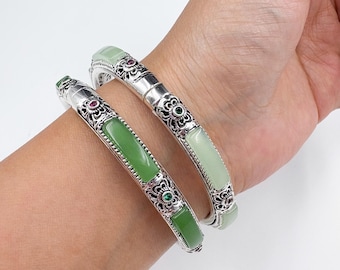 May include: Two silver bangles with green jade stones and ornate floral designs. The bangles are on a wrist, showcasing the contrast between the polished silver and the vibrant green of the jade. The bangles have red and green accents.