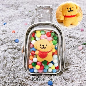 May include: A clear sling bag with a zip, filled with colourful pom-poms and a yellow dog toy wearing a red heart. The bag has a white trim and the text "Butter Dog" at the bottom.