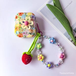 K&ouml;nnte beinhalten: Eine wei&szlig;e AirPods-H&uuml;lle mit Blumenmuster, einer gro&szlig;en gelben Blume und einem rosa Zentrum. Ein roter geh&auml;kelter Erdbeer-Anh&auml;nger und eine Blumen-Handgelenkschlaufe mit verschiedenen farbigen Blumen und durchsichtigen Perlen sind befestigt.