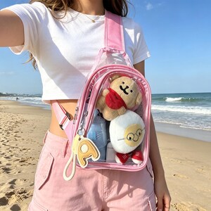 Op de afbeelding: Een doorzichtige, roze schoudertas met een teddybeer en een pluchen figuur erin. De tas heeft een roze band en een goudkleurige letter "A" sleutelhanger. De tas wordt op een zonnige dag op het strand over de schouder gedragen.