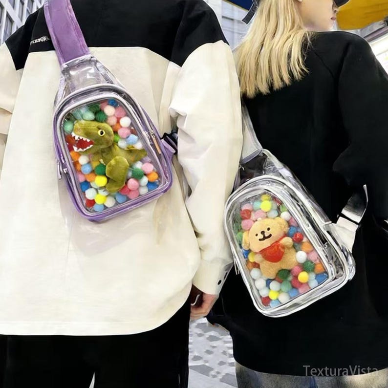 Puede incluir: Bolsos cruzados transparentes con correa morada, llenos de juguetes y pompones de colores. Uno contiene un dinosaurio verde, el otro un osito de peluche. Ambos se llevan cruzados.